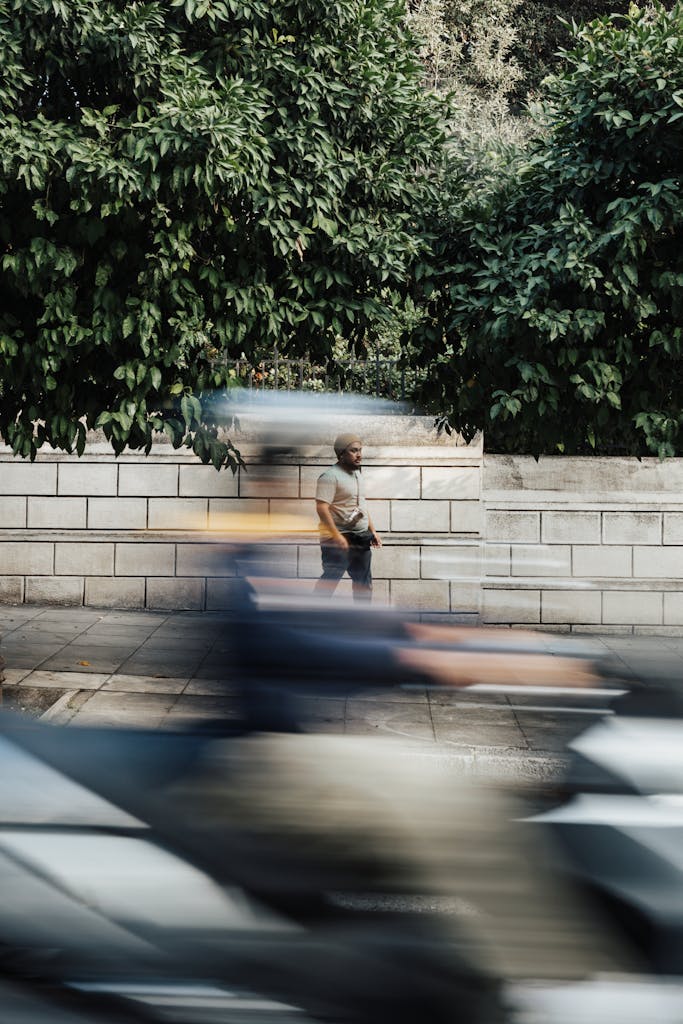 A scooter speeds past, blurring in motion, while a man stands in focus against a leafy wall.